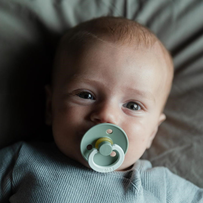 A light-haired baby in a gray shirt lies down, eyes on the camera. They use a BIBS Natural Latex Rubber Glow in the Dark Pacifier, Size 1 (0-6 Months), for comfort and safety. The background is soft and neutral-colored.
