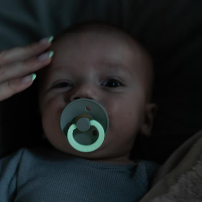 A baby with a BIBS Natural Latex Rubber Glow in the Dark Pacifier, Size 1 (0-6 Months) in their mouth lies down as a hand with painted nails gently touches their forehead. The dim lighting creates a calm and soothing atmosphere.