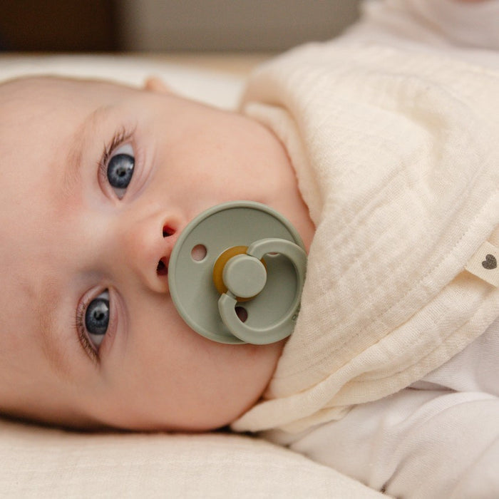 A baby lies down wearing a white garment and cream bib, with wide blue eyes gazing upward and a Natural Latex Rubber Pacifier by BIBS, Size 1 (0-6 Months), BPA-free, in their mouth.