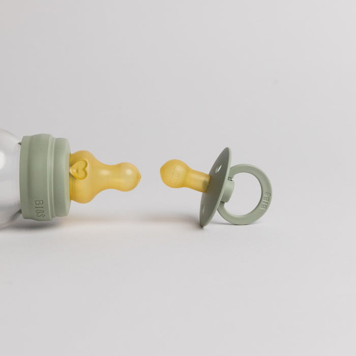 A BPA-free green baby bottle with a yellow nipple lies on its side beside a BIBS Natural Latex Rubber Pacifier, Size 1 (0–6 months), in matching colors, standing upright. Both are set against a plain white background, offering reliable breastfeeding support.