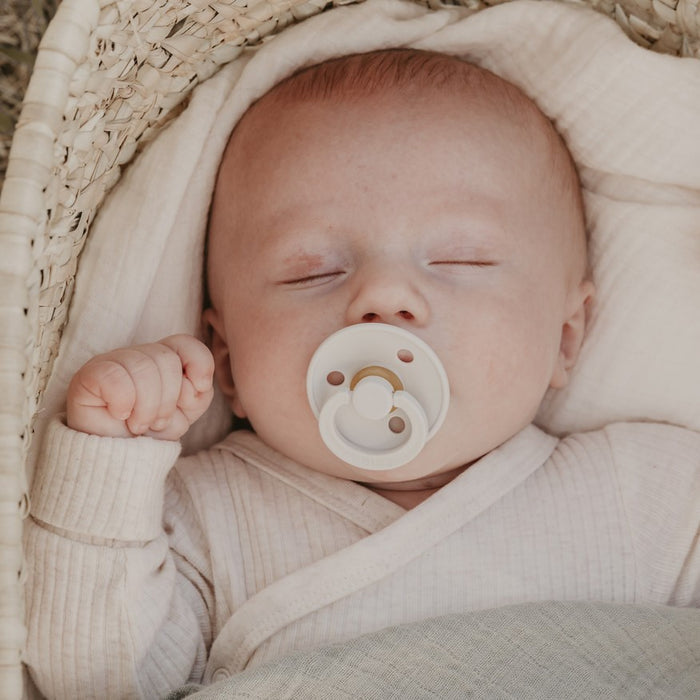 A baby sleeps peacefully in a woven basket, wearing a cream outfit and holding a BIBS Natural Latex Rubber Pacifier, Size 2 (6-18 Months) in its mouth. The BPA-free pacifier complements the babys light blanket, offering comfort as they rest with one hand gently clenched.