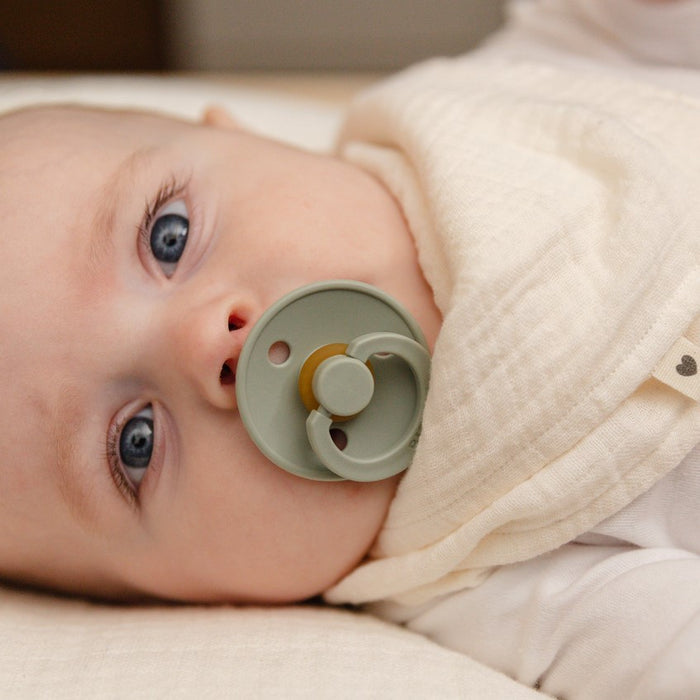 A baby with blue eyes contentedly sucks on a green BIBS Natural Latex Rubber Pacifier, Size 2 (6-18 Months), while dressed in a soft white outfit and partially covered by a cream-colored blanket, creating a serene scene of comfort and gentle breastfeeding support.