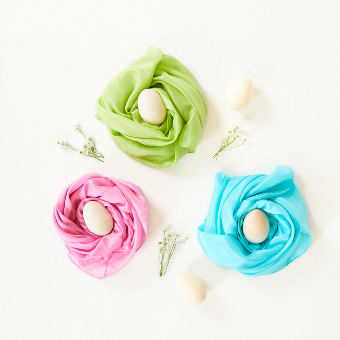 Three pastel-colored scarves from Sarahs Silks Natural, Non-Toxic Easter Egg + Playsilk Dye Kit wrap around eggs with a nest-like look, accented by small white flowers. Two extra eggs on the white backdrop highlight colors made with organic fruit and vegetable extracts.