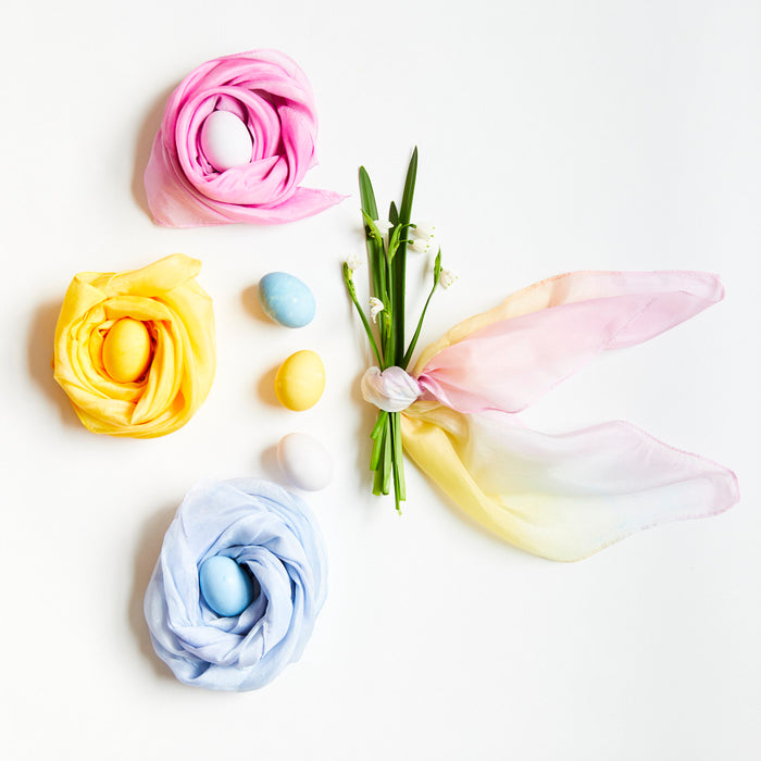 On a white background, colorful eggs wrapped in pink, yellow, and blue silk scarves from Sarahs Silks Natural, Non-Toxic Easter Egg + Playsilk Dye Kit are displayed. Two loose scarves embrace dyed eggs, while a bunch of white flowers is tied with a multicolored scarf nearby.