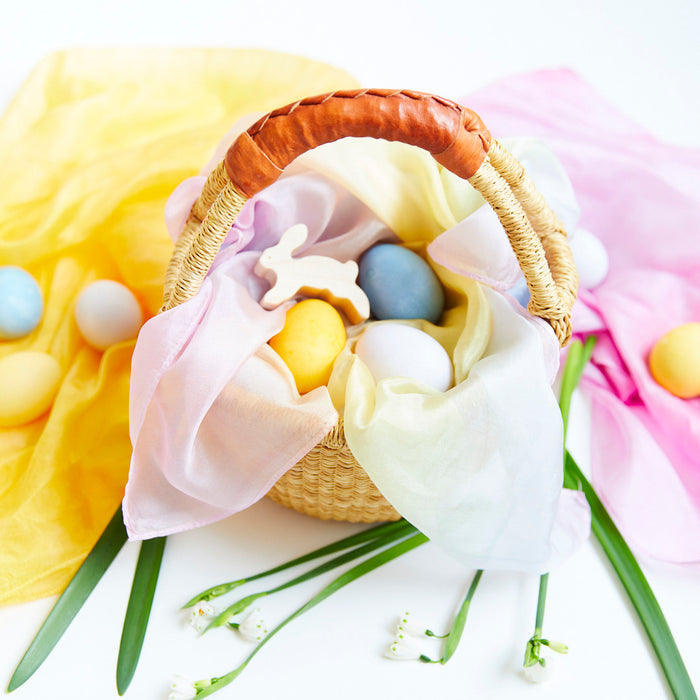 A wicker basket filled with colorful Easter eggs created using Sarahs Silks Natural, Non-Toxic Easter Egg + Playsilk Dye Kit, pastel fabric, and a bunny-shaped cookie. Surrounded by draped pastel cloths and green leaves on a white background.