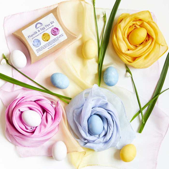 An Easter egg display showcases fabric roses in pink, yellow, and blue. Eggs, dyed with Sarahs Silks Natural, Non-Toxic Easter Egg + Playsilk Dye Kit, nestle inside. Green leaf accents complete the design against a vibrant backdrop for a festive touch.