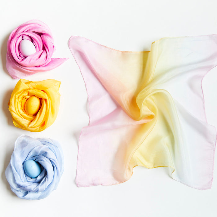 Three eggs wrapped in vibrant pink, yellow, and blue silk scarves from the Natural, Non-Toxic Easter Egg + Playsilk Dye Kit by Sarahs Silks are arranged on the left. A thin scarf dyed with organic fruit and vegetable extracts is spread loosely on the right against a white background.