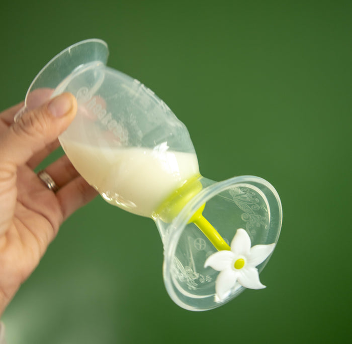 A hand holds an upside-down transparent silicone cup filled with milk, featuring a yellow straw and white flower-shaped valve. The solid green backdrop highlights the innovative design, matching Haakaa’s New Mom Starter Pack for efficient breastfeeding accessories.