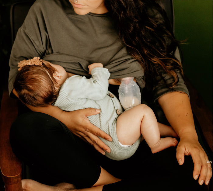 A woman with long dark hair, dressed in dark clothing, sits in a chair breastfeeding her infant who is in a light green outfit. Nearby are a milk bottle and Haakaas New Mom Starter Pack disposable breast pads as she calmly nurtures her baby.