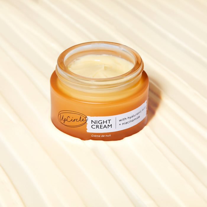 An open jar of UpCircles Night Cream with Hyaluronic Acid + Niacinamide, featuring blueberry extract, rests on a light textured surface. The pale cream in an amber jar offers enriched nighttime nourishment.