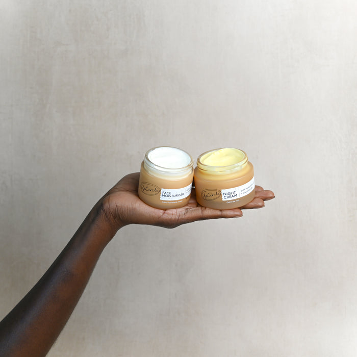 A hand showcases two small skincare jars against a textured beige backdrop. The Day Moisturizer with white cream sits alongside the UpCircle Night Cream with Hyaluronic Acid + Niacinamide, enhanced for radiant skin.