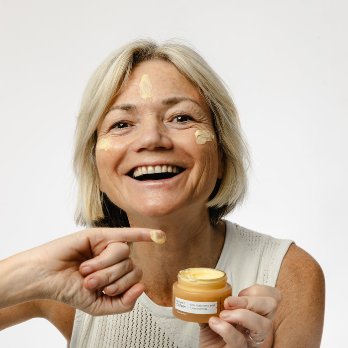 A grey-haired woman smiles as she applies UpCircles Night Cream with Hyaluronic Acid + Niacinamide, infused with blueberry extract, to her face using a finger. She wears a sleeveless white top and holds a small jar of yellow cream against a plain white background.