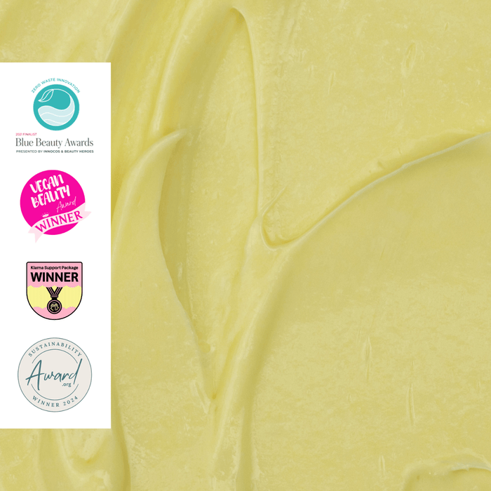 Close-up of a creamy textured yellow substance with several award logos on the left side. The awards include "Blue Beauty Awards," "Global Green Beauty Awards," "Mama &amp; Baby Awards," and "Beauty Shortlist Awards," highlighting recognition for quality and sustainability.