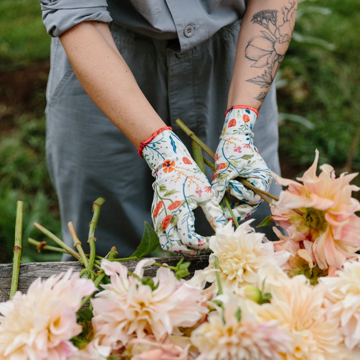 Wearing Nitrile Weeder Gloves in the Garden of Paradise print by Womanswork, a person with a floral tattoo on one arm tends to large pale pink dahlias. They are dressed in grey and work outdoors against a blurred green backdrop.