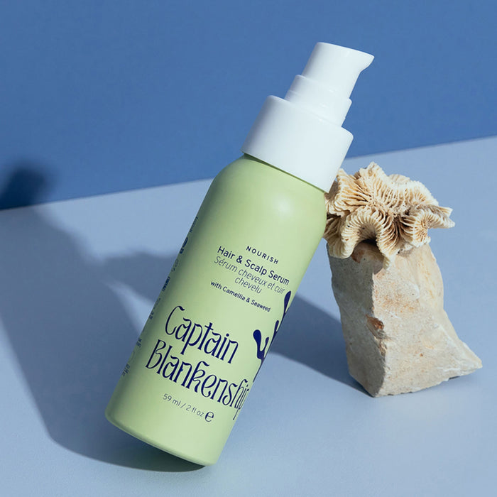 Displayed on a light blue surface with a textured stone and coral, Captain Blankenships Nourish Hair & Scalp Serum in a green bottle and pump dispenser is highlighted against a soft blue background. Featuring nourishing serum, its enriched with organic botanical oils.