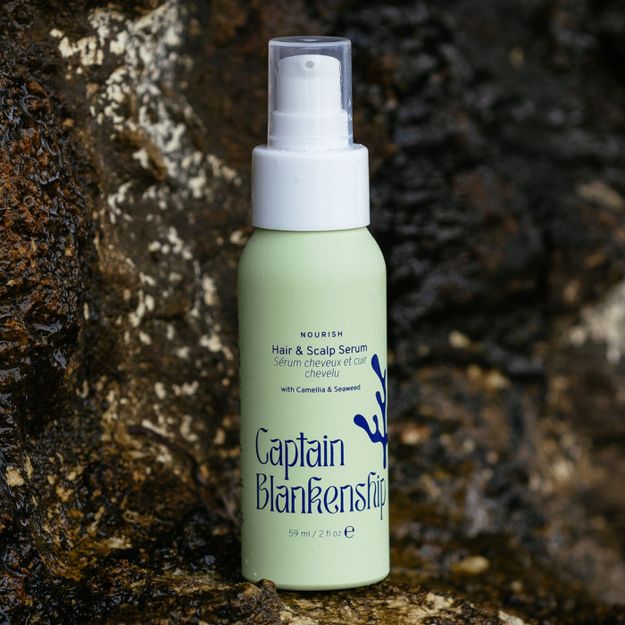 A green bottle of Captain Blankenships Nourish Hair & Scalp Serum, featuring a spray pump and enriched with camellia and seaweed, is photographed against a textured rock. This 59ml serum boasts organic botanical oils for effective hair care.
