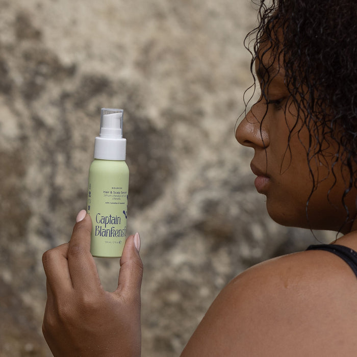 A curly-haired individual holds a green bottle of Captain Blankenships Nourish Hair & Scalp Serum, enriched with organic botanical oils. A rocky background complements the seaweed-powered hair care as they admire the nourishing serum in profile.
