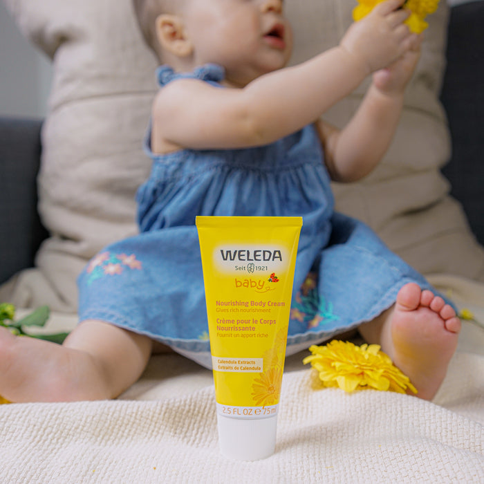 A baby in a blue dress sits on a couch holding a yellow flower. In the foreground, Weledas Nourishing Body Cream is placed on a white surface, ideal for nurturing babys skin.