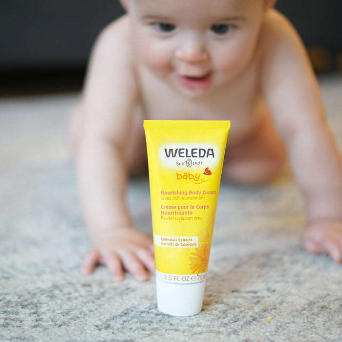 A baby crawls on the carpeted floor, captivated by a yellow tube of Weledas Nourishing Body Cream with calendula extract, ideal for nourishing babys skin. Nearby, a TerraCycle recycling program reminder ensures each tube finds new life beyond its use.