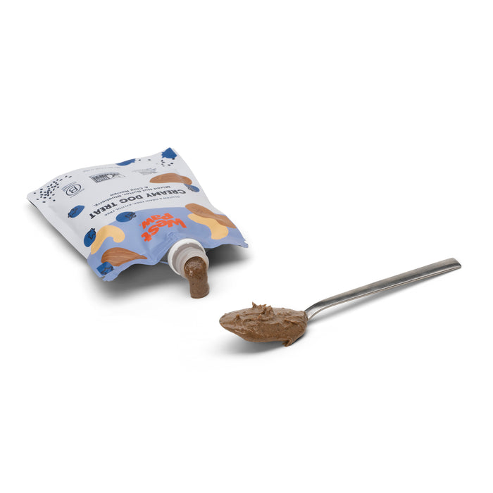 A pouch of West Paws Nut Butter Blueberry Creamy Dog Treat lies on its side, paste oozing from the mess-free dispenser. Nearby, a spoon holds a dollop, while the design features bones and playful text.