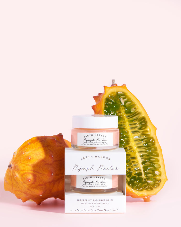 The NYMPH NECTAR Superfruit Radiance Balm by Earth Harbor is elegantly showcased in front of two kiwano fruits, one whole and one sliced, against a light pink background. This luxurious balm features a vegan wax-free formula to enrich your skin while you sleep.