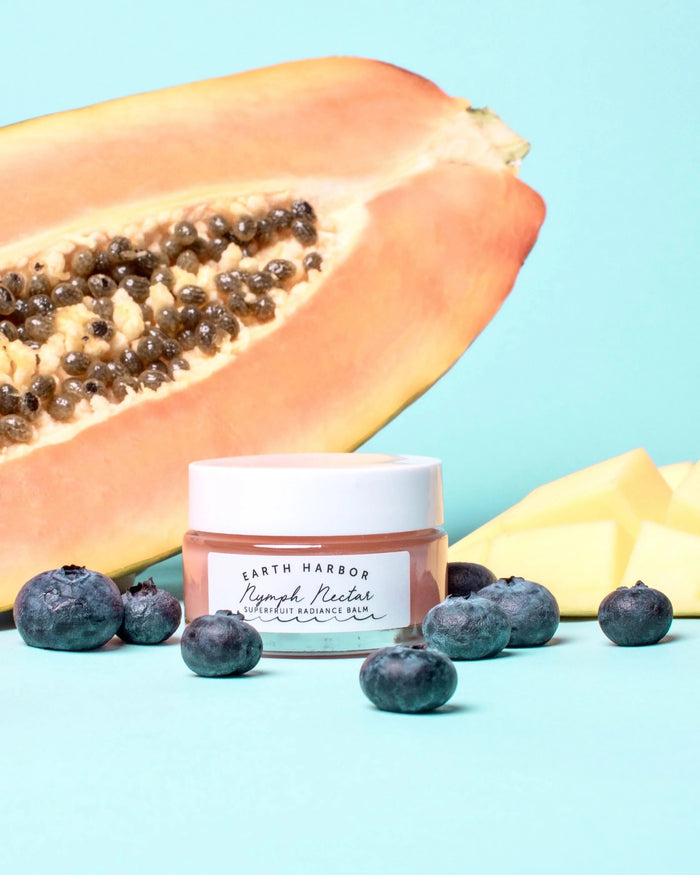 Amid halved papaya and ripe pieces on a light blue backdrop, sits Earth Harbor labeled skincare jar of NYMPH NECTAR Superfruit Radiance Balm. Blueberries accent this vibrant scene featuring a luxurious vegan wax-free formula.