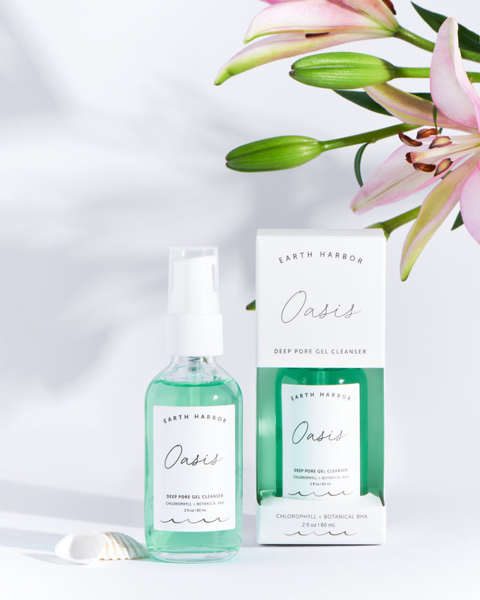 A clear bottle of Earth Harbors OASIS Deep Pore Gel Cleanser with a spray nozzle is enriched with purifying botanicals and stands next to its white and green packaging. The scene features pink lilies and a small shell against a light backdrop, perfect for sensitive skin.