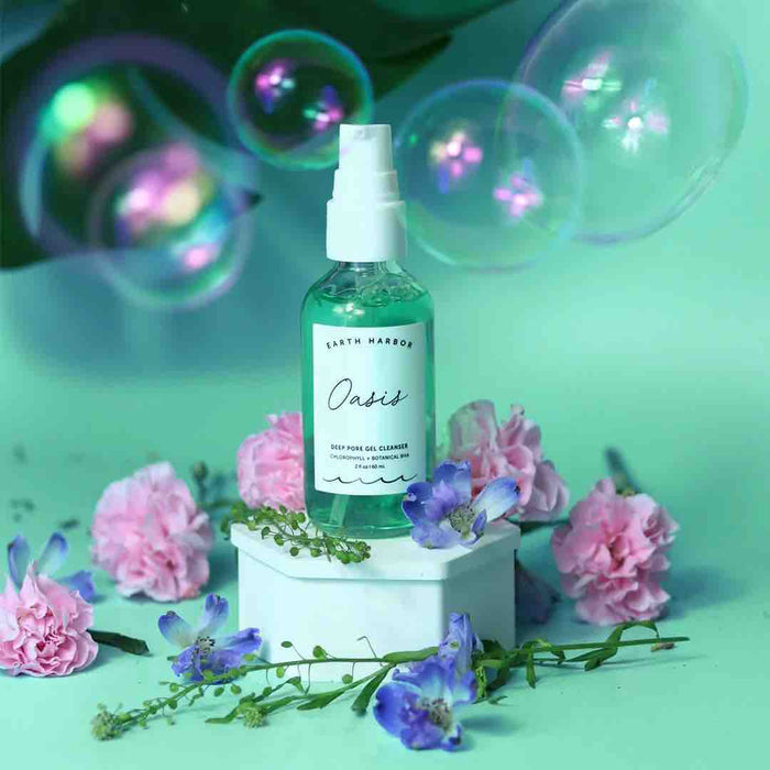 Earth Harbors OASIS Deep Pore Gel Cleanser is showcased on a white pedestal with pink carnations and purple flowers, perfect for sensitive skin. Its purifying botanicals gleam against a mint green backdrop, enhanced by bubbles and leafy greens for an ethereal effect.