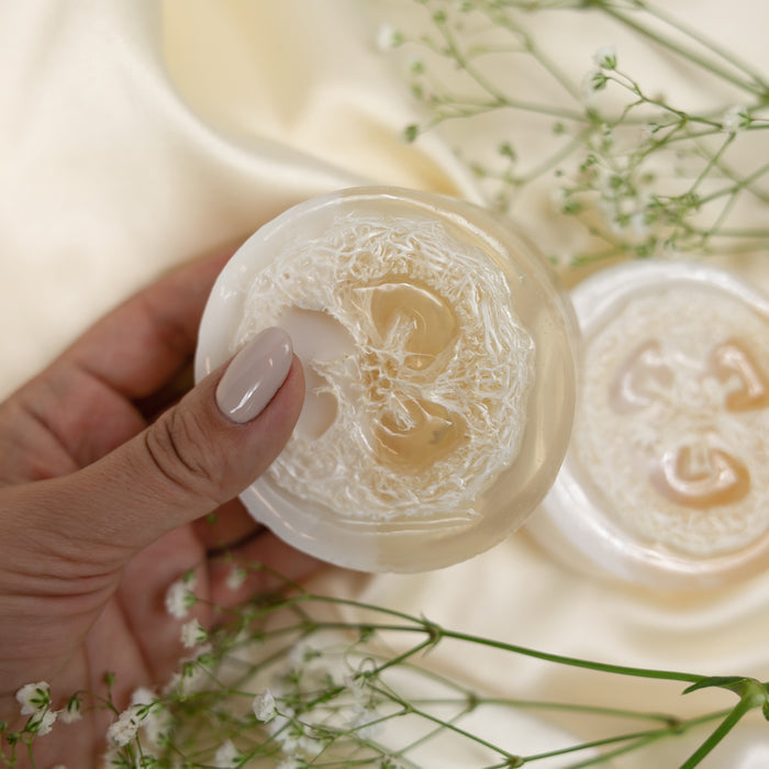 A hand with manicured nails holds a transparent Soapymania Oatmeal, Milk & Honey Loofah soap embedded with fibers. In the background, another similar soap and delicate white flowers rest on soft, light fabric.