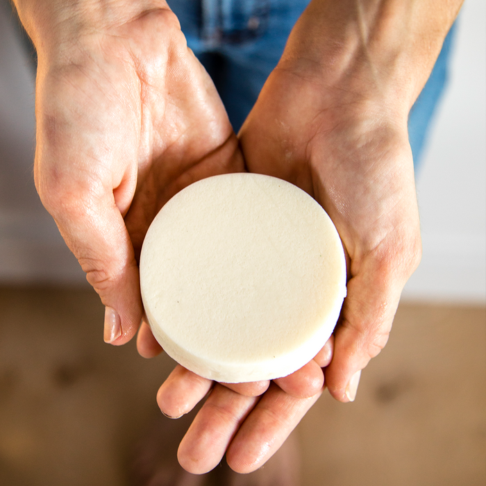 Hands gently cradle the Spinster Sisters Co. Oatmeal Milk & Honey Body Butter Bar, infused with Fair Trade shea butter. The neutral, softly focused background highlights this vegan skincare product resting on a light surface.