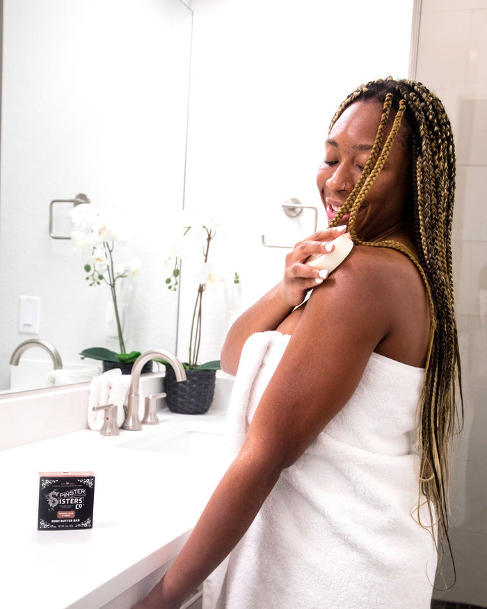 With long braided hair wrapped in a towel, a woman smiles as she uses an Oatmeal Milk & Honey Body Butter Bar on her shoulder. In the bright bathroom with a white countertop and faucet, a Spinster Sisters Co. box sits beside her, and white flowers adorn the background. The bar features Fair Trade shea butter.