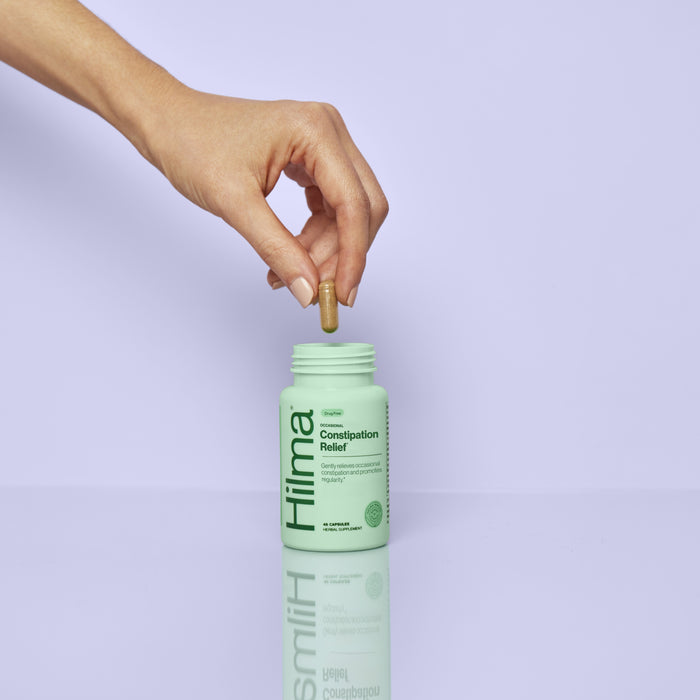 A hand holds a capsule above an open green bottle labeled Hilma Gentle Bowel Movement Support on a smooth light purple surface with a matching background.