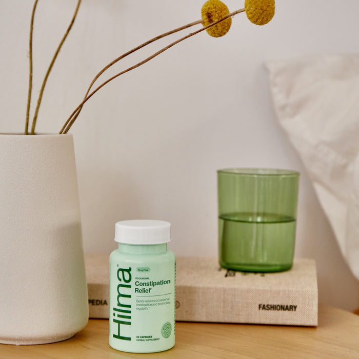 A bottle of Hilma Gentle Bowel Movement Support sits on a nightstand beside a beige vase with yellow flowers, a green glass, and a book showing part of the title FASHIONARY.