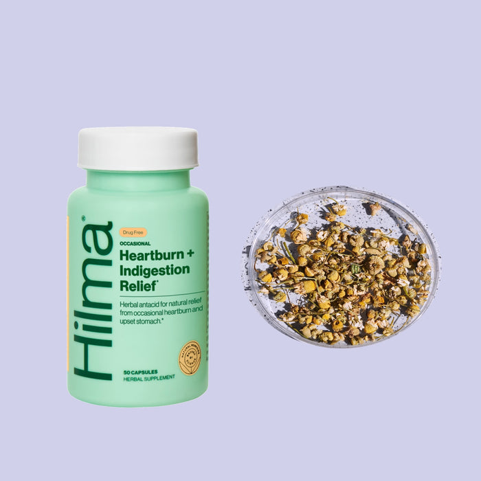 A green bottle labeled Hilma Occasional Heartburn + Indigestion Relief with 50 capsules sits beside a small bowl of dried chamomile flowers, promising digestive comfort against a light purple background.
