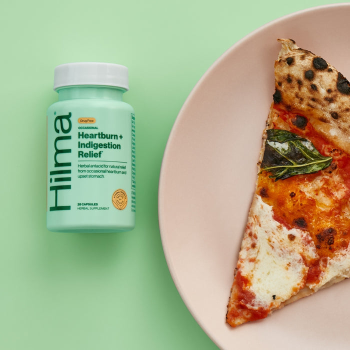 A bottle of Hilma Occasional Heartburn + Indigestion Relief rests beside a cheesy, basil-topped pizza slice on a light green background, offering digestive comfort for any post-pizza heartburn.