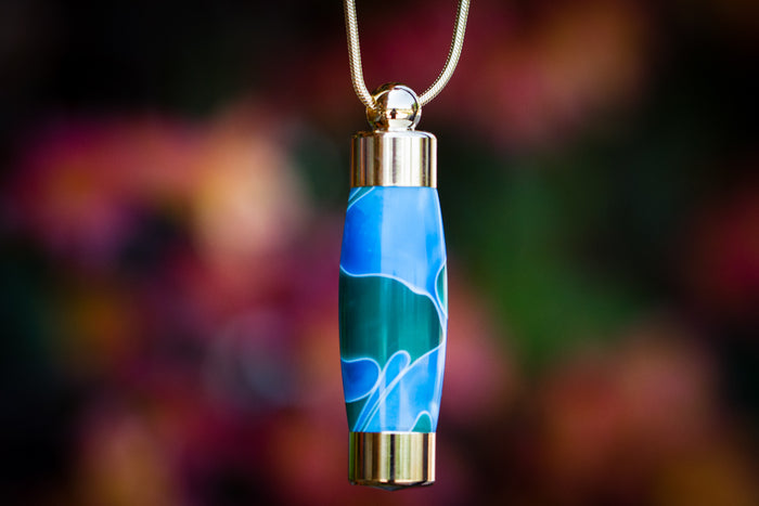 Ocean blue teal marbled keepsake pendant on a gold chain against a colorful blurred background.