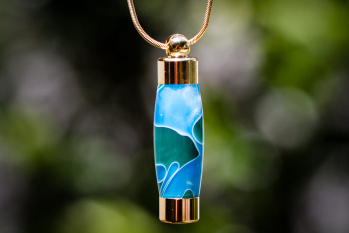Close-up of ocean blue acrylic keepsake urn pendant with gold caps on a gold chain against a blurred background