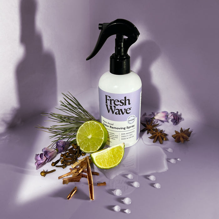 A bottle of Fresh Wave Odor Removing Fabric and Linen Spray rests on a purple surface, adorned with pine needles, lime slices, cloves, cinnamon sticks, and star anise. It is designed to naturally refresh fabrics and rooms, with white droplets accentuating the scene.