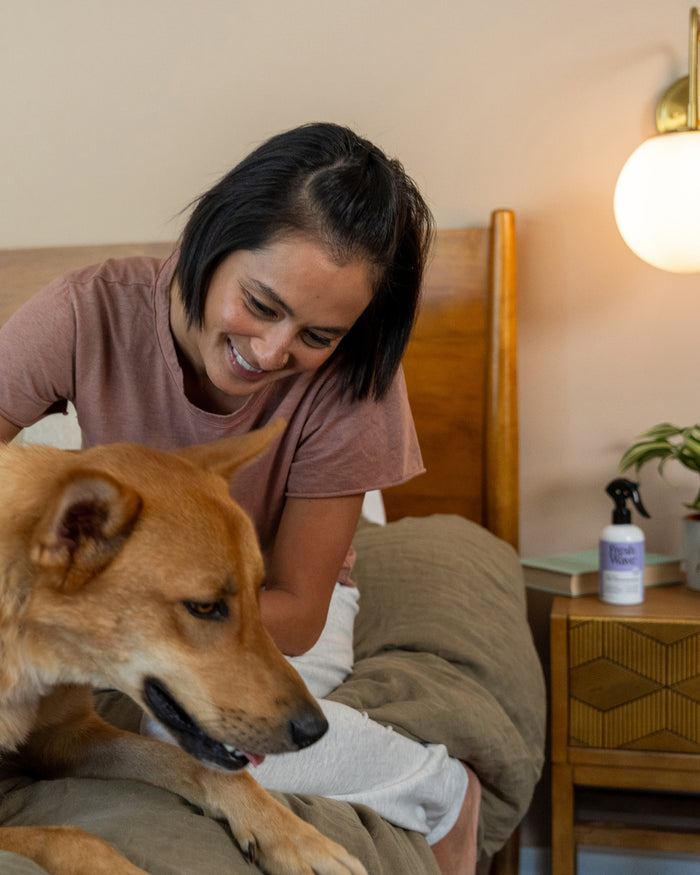 A person with short dark hair, in a brown shirt, smiles on a bed while petting a large tan dog beside them. In the background sits a wooden bedside table with Fresh Waves Odor Removing Fabric and Linen Spray and a lamp, perfect for instantly refreshing fabrics and rooms.