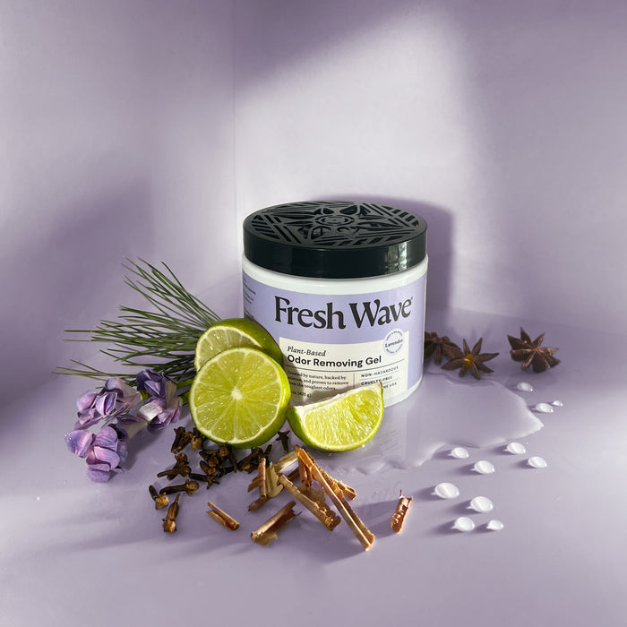 A Fresh Wave Odor Removing Gel container, infused with natural plant oils for continuous odor control, is placed among lime slices, pine needles, star anise, purple flowers, cloves, and cinnamon sticks. White liquid drops are scattered on a light purple surface.