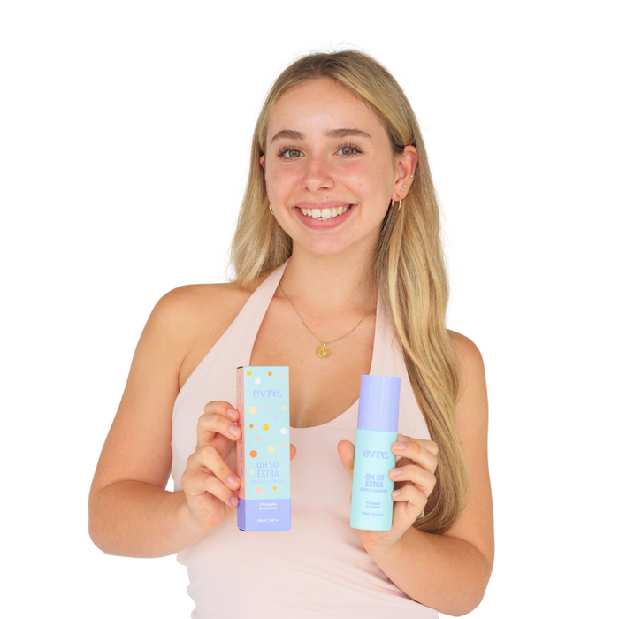 A smiling woman with long blonde hair holds an evre. Oh So Extra Pineapple & Coconut Face Cleanser box and matching cleanser bottle in pastel blue and purple packaging. She wears a light pink top, standing against a white background.
