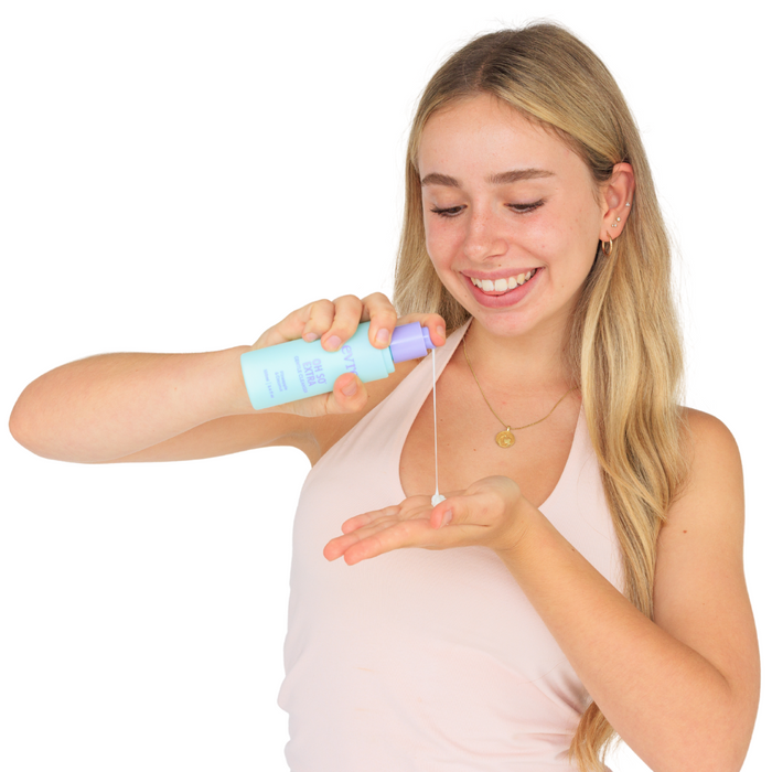 A person with long blonde hair and a pink tank top smiles while dispensing evre.s Oh So Extra Pineapple & Coconut Face Cleanser from a blue and purple bottle onto their palm. The background is plain white.