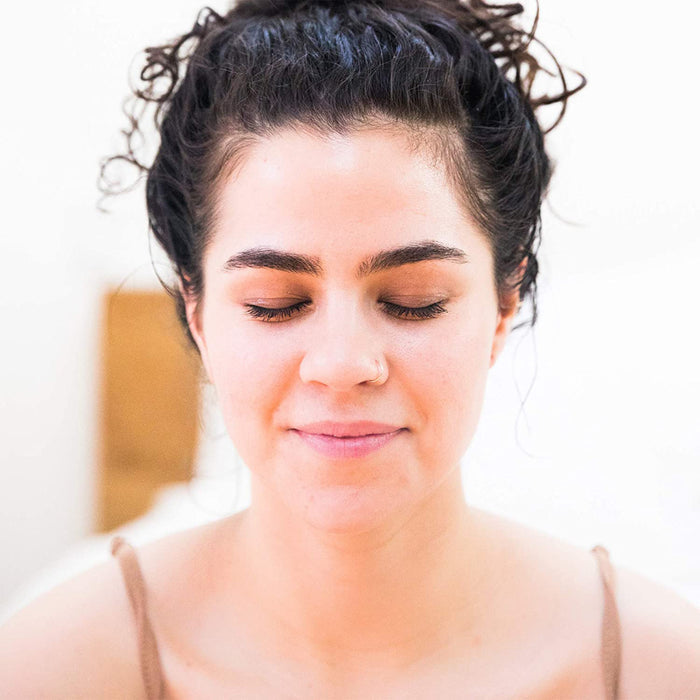A curly-haired woman with closed eyes and a slight smile glows while wearing a light top, thanks to Beauty by Earths Oil Control Facial Moisturizer with natural ingredients. Her calm expression merges seamlessly into the softly blurred, warm-toned background.
