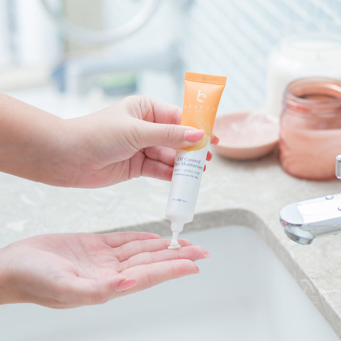 In a bathroom with a marble countertop, a person dispenses Beauty by Earths Oil Control Facial Moisturizer from an orange and white tube onto their palm. A copper jar and white candle in the blurred background set the tone for this Natural Ingredients skincare ritual.