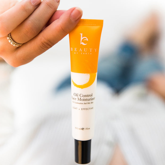 A hand holds a tube labeled Beauty by Earth Oil Control Facial Moisturizer, featuring organic aloe vera. The yellow and white design emphasizes its suitability for oily and combination skin, against a softly blurred background.