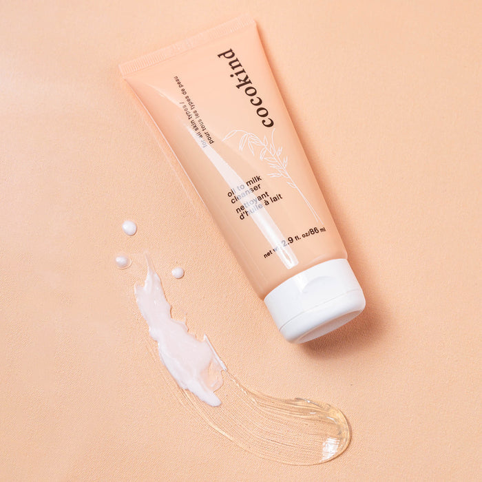 A tube of Cocokinds Oil to Milk Cleanser, enriched with an organic oil blend, rests on a light peach surface. The packaging features white text and illustrations. Beside it, a dollop highlights its creamy, oil-like texture.