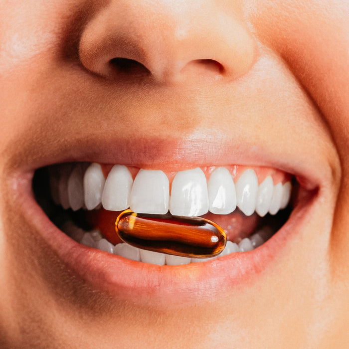 A close-up of a smiling mouth with white teeth gently holding a transparent, amber Omega-3 Fish Oil capsule from True Grace between the teeth.