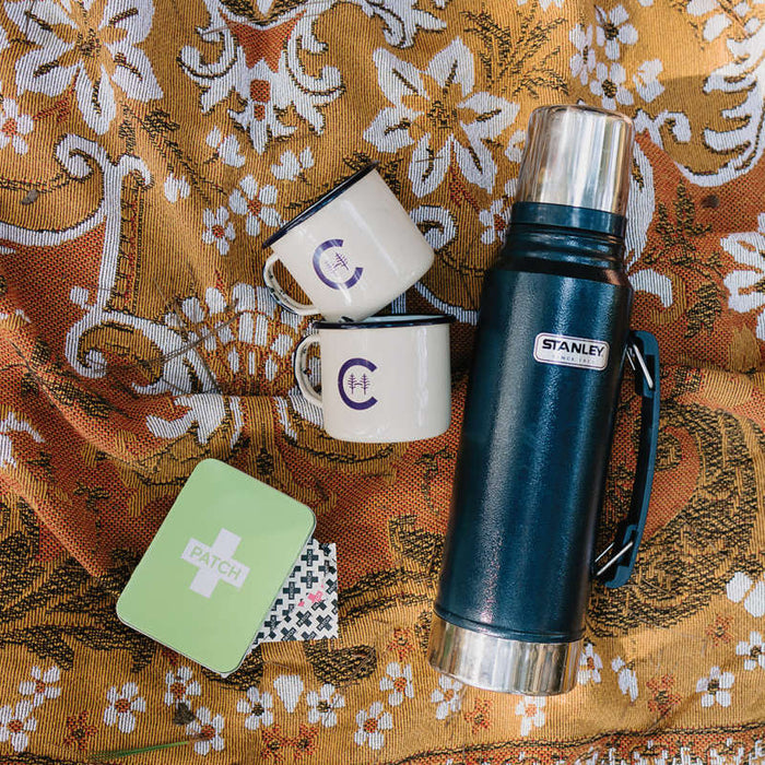 A vintage thermos and two enamel mugs with a C logo sit on a patterned fabric, next to the PATCH On-The-Go Travel Kits green tin with a cross symbol, hinting at an included first aid kit, creating a relaxed outdoor picnic vibe.