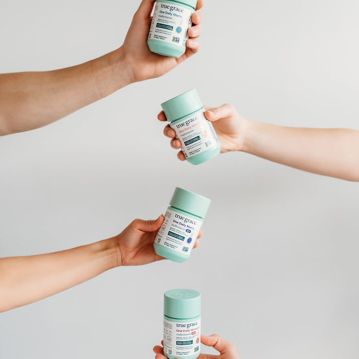 Four hands each hold a container of True Grace dietary supplements against a light background. Each container is designed with a pastel green lid and a white label with blue and black text.