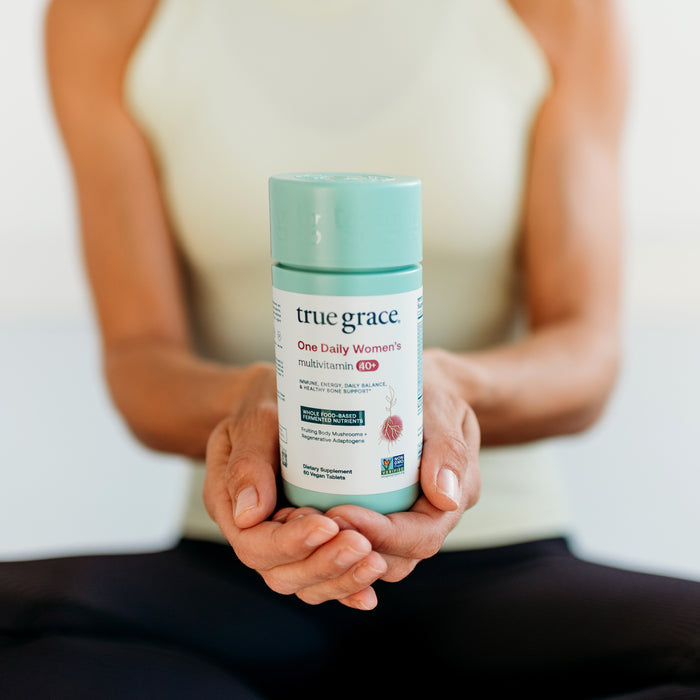 A person in a light tank top holds a light green bottle of "true grace One Daily Women's multivitamin 40+." The bottle has pink graphics and is designed for women's health. The person is sitting cross-legged.